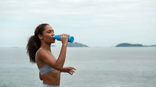 Active woman drinking from a blue water bottle by the ocean, replenishing electrolytes after exercise.