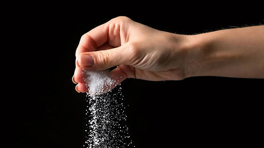 A hand pinching and sprinkling a large amount of white salt against a black background, symbolizing the excessive sodium content in salty-tasting electrolyte products.